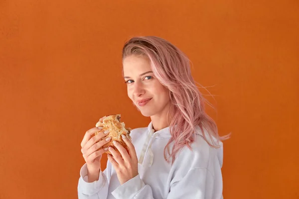 Mladá žena s růžovými vlasy se podívá do kamery a drží v ruce hamburger. — Stock fotografie