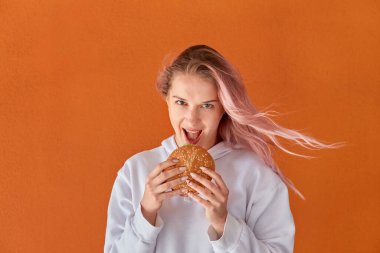 Bir kadın büyük lezzetli bir hamburger yer ve kameraya bakar. Turuncu arkaplan.