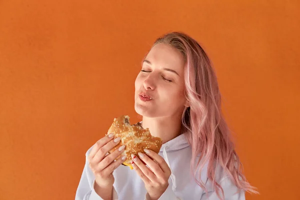 Mladá žena s růžovými vlasy jí rychlé občerstvení s chutí. oranžové pozadí. — Stock fotografie