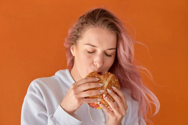 Hladová mladá žena s růžovými vlasy a jíst velký burger s chutí k jídlu. — Stock fotografie