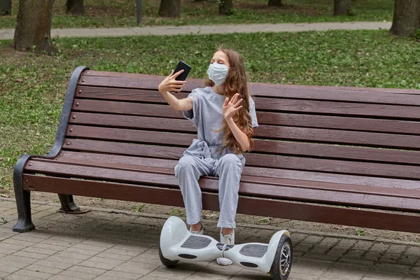 Uma menina em uma máscara leva uma selfie em um banco, seus pés estão em um giroscópio. — Fotografia de Stock