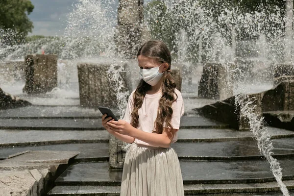 Uma adolescente em uma máscara médica em um parque com um celular nas mãos. — Fotografia de Stock