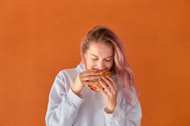 Aç genç bir kadın iştahlı büyük bir burger yer. Turuncu arkaplan.