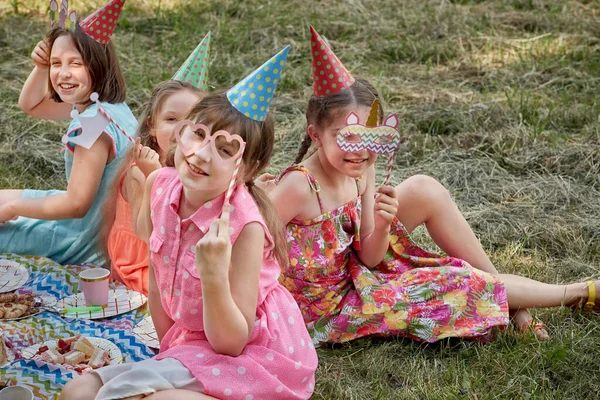 Dziewczyny świętują w maskach karnawałowych, kapeluszach. Zabawa piknik dla dzieci z przyjaciółmi. — Zdjęcie stockowe