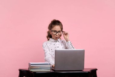 Pembe arka planda oturan kız şaşırmış yüzlü bir bilgisayarla masada oturuyor.