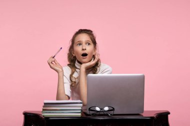 Şaşırmış yüzlü kız öğrenci elinde kalemle dizüstü bilgisayarda oturuyor.