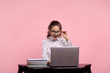 Masanın üzerinde bir dizüstü bilgisayar ve kitaplar var. Gözlüklü kız masada oturuyor..