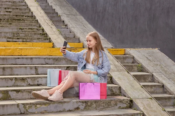 Симпатичная девушка делает селфи сидя на лестнице с покупками в красочных сумках. — стоковое фото