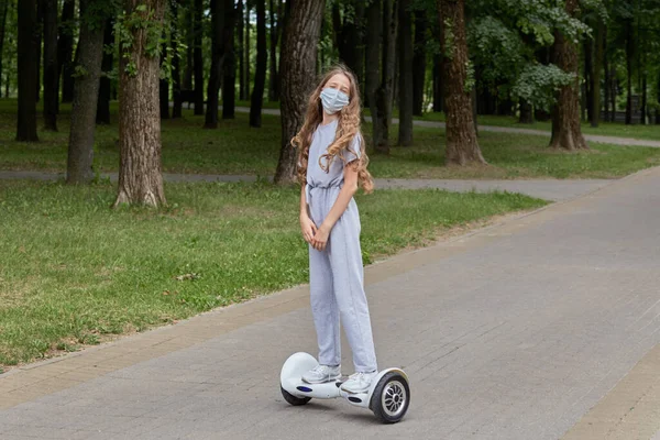 Uma menina monta uma scooter giroscópio, prancha em uma máscara médica no verão. — Fotografia de Stock