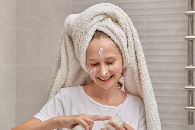 Kafasında havlu olan bir kız yüzüne beyaz nemlendirici sürüyor..