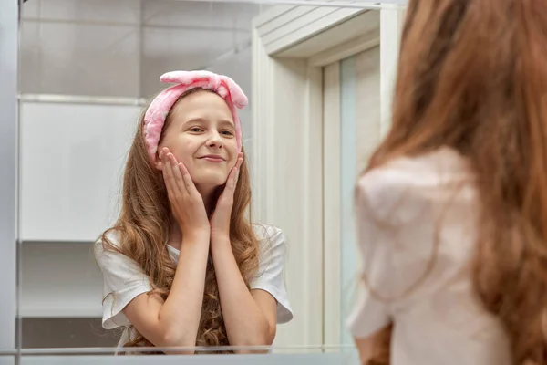 Girl with long blonde hair teen looks in the mirror. Pink headband on her head.