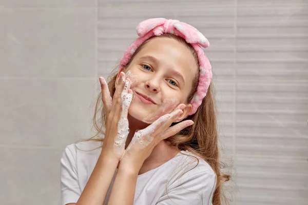 Beautiful young blonde girl washes her face with foam in the bathroom.