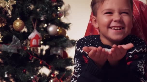 Leuke gelukkige jongen houdt kunstmatige sneeuw in zijn hand en blaast ze in de camera. — Stockvideo