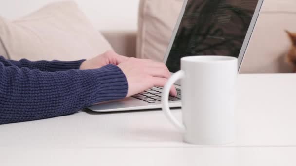 En la mesa hay una taza de café y un hombre trabajando en línea en casa en un ordenador portátil. — Vídeos de Stock