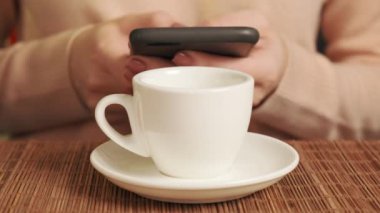 Elinde cep telefonu olan ve beyaz bardakta kahve içen bir kadın..
