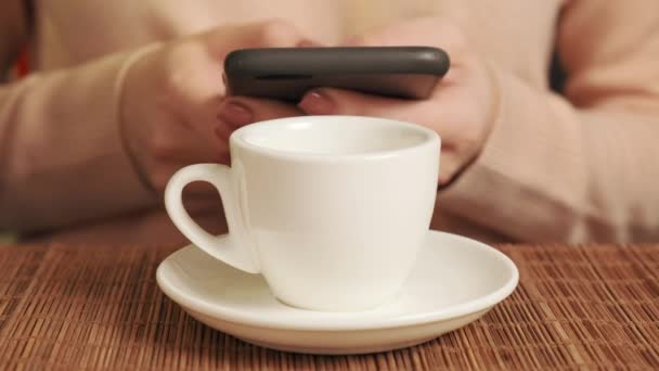 Una mujer con un teléfono móvil en sus manos y tomando café en una taza blanca. — Vídeos de Stock