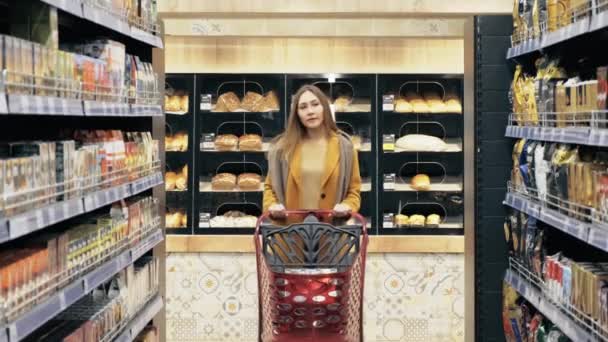 Una mujer camina a través de una tienda al por menor con un carrito de compras y selecciona productos. — Vídeo de stock