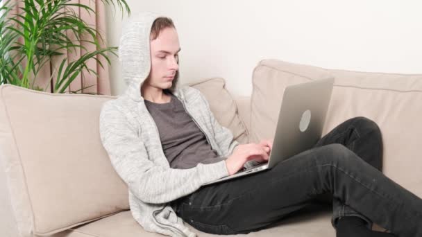 Młody mężczyzna pracuje w domu na kanapie i pije gorącą kawę w białym kubku.. — Wideo stockowe