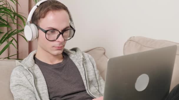 Un hombre con gafas para la visión trabaja con auriculares tumbados en el sofá en una computadora portátil. — Vídeos de Stock