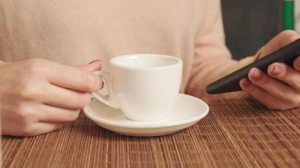 Una mujer compra regalos en línea en su teléfono y bebe café fragante en una taza. — Vídeos de Stock