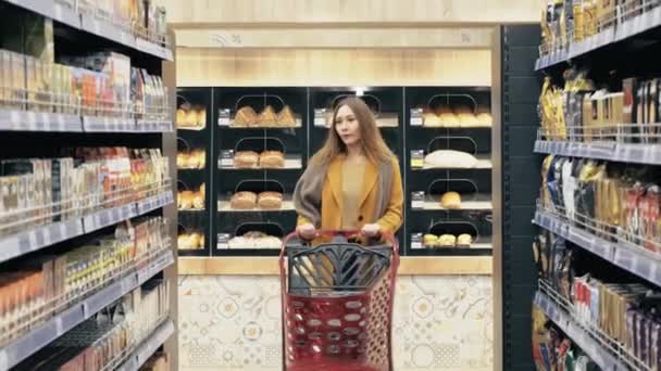 En ung kvinna går med en kundvagn och väljer produkter i butiken. — Stockvideo