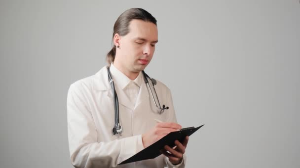 Een mannelijke arts met lang haar in een medisch gewaad schrijft op papier op een klembord. — Stockvideo
