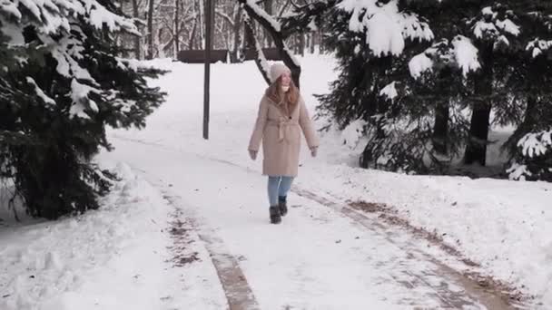 베이지 코트를 입은 젊고 아름다운 금발의 여성이 눈덮인 도로를 걷고 있습니다. — 비디오