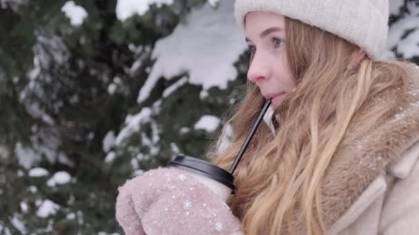 Uma mulher bebe uma bebida de um copo e uma palha no inverno frio e sorri. — Vídeo de Stock