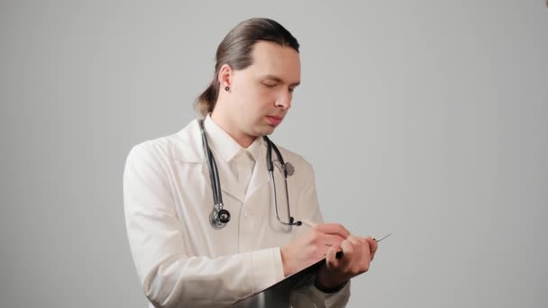 Een jonge mannelijke arts in een medisch gewaad schrijft op papier op een klembord. — Stockvideo