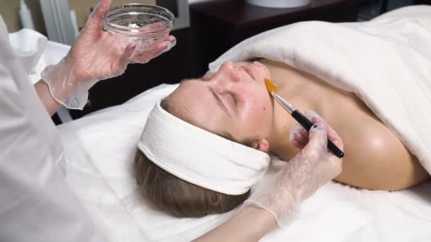 Kosmetologen applicerar en transparent mask med en borste på klientens ansikte. — Stockvideo