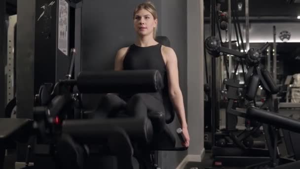 A woman performs exercises sitting on a simulator for athletic legs and buttocks — Stock Video