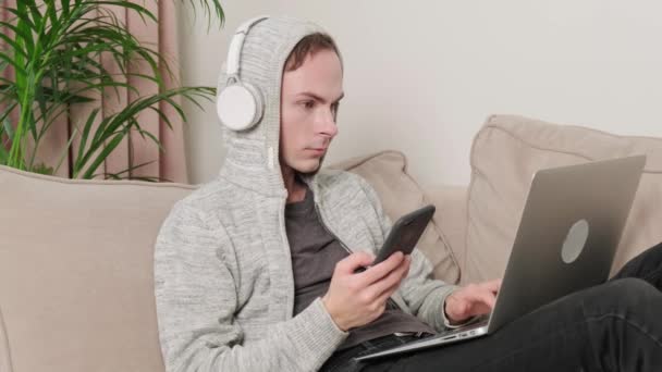 彼の手に携帯電話を持つ男はソファの上に横たわるヘッドフォンで動作します. — ストック動画