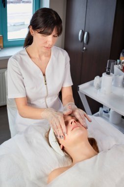 Kozmetik uzmanı genç bir kadın eldiven takan bir müşteriye masaj yapıyor..