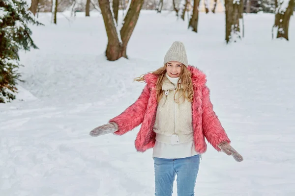 Młoda kobieta przechadza się po zimowym parku w różowym futrze i dzianym kapeluszu. — Zdjęcie stockowe