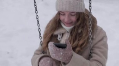 Bir kadın kışın salıncakta sallanır ve elinde akıllı bir telefonla konuşur..