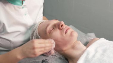 Bir kozmetolojistin elleri pamuklu genç bir adamın yüzünü temizler..