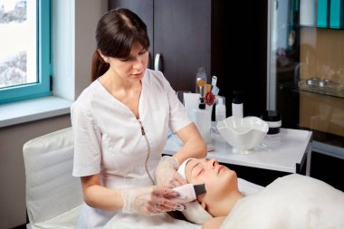 Tıbbi önlük giyen bir kozmetik uzmanı bir müşteri için yüz temizleme prosedürü uygular.