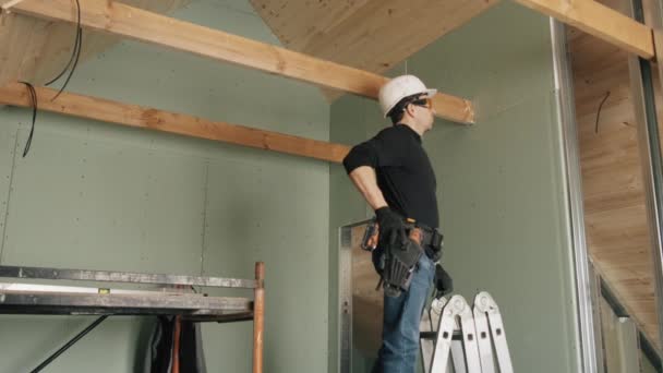 Un trabajador de la construcción se para en una escalera y trabaja con un taladro. — Vídeo de stock
