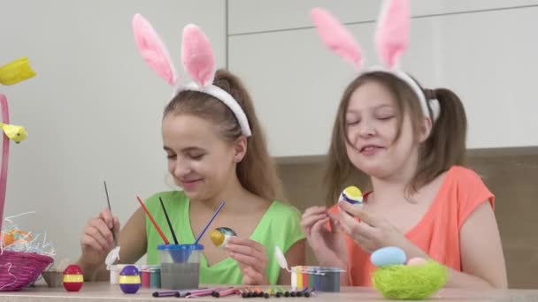 Dos niñas decoran los huevos de Pascua con un cepillo y se ayudan mutuamente, hablar y reír. — Vídeos de Stock