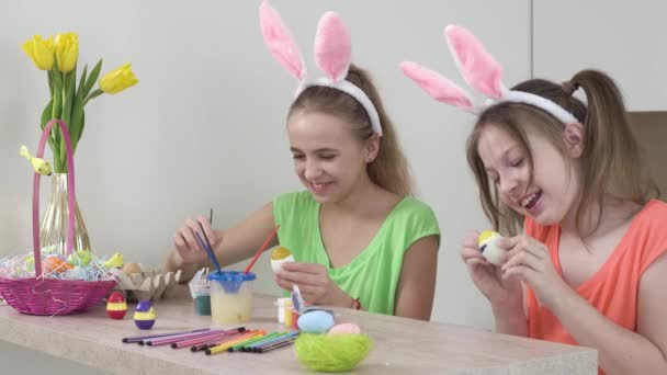 Las niñas están decorando huevos de Pascua y riendo, con orejas de conejo en la cabeza. — Vídeos de Stock