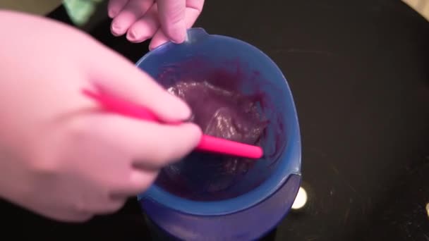 Руки в розовых латексных перчатках смешивают фиолетовый краситель для волос в пластиковом контейнере. — стоковое видео