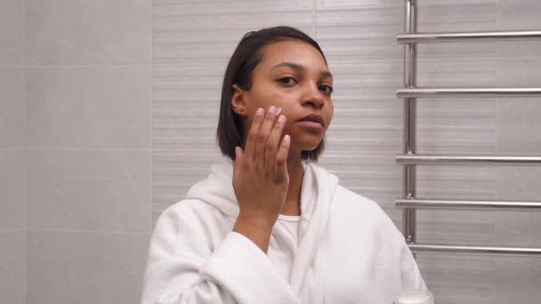 Жінка з темною шкірою використовує зволожуючий крем для обличчя вранці у ванній . — стокове відео