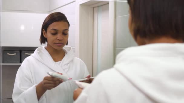 Een vrouw kijkt in de spiegel en poetst haar tanden met tandpasta, een tandenborstel. — Stockvideo