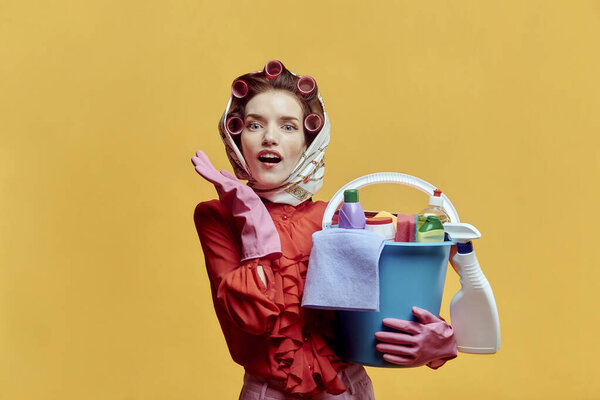 A young surprised female cleaner holds a plastic bucket with cleaning tools.