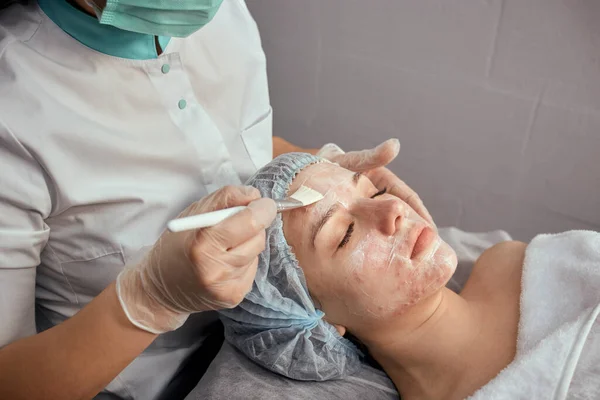 A female cosmetologist in a medical mask applies cream to the client face. — Stock Photo, Image