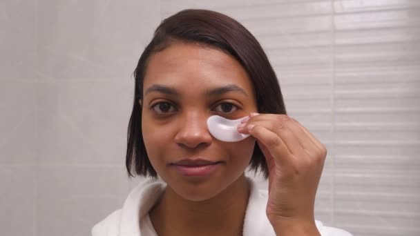 A young woman with dark skin uses white patches for the eyes, against wrinkles. — Stock Video