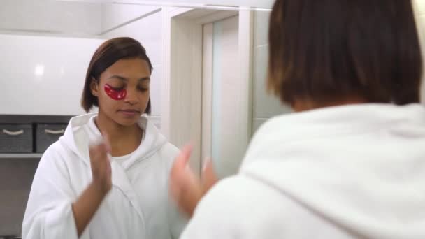 Egy fiatal, gyönyörű nő belenéz a tükörbe, és szemkötőt használ. Spa nap. — Stock videók
