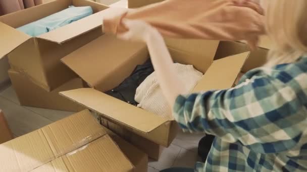 Una mujer con una camisa a cuadros pone su ropa en cajas de cartón. — Vídeos de Stock