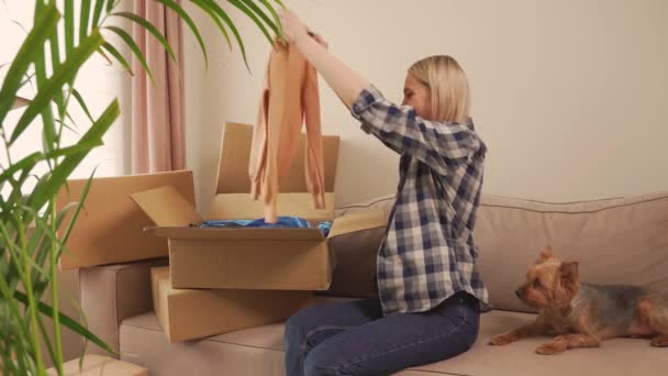 Una mujer sentada en el sofá abre una caja de ropa después de mudarse. Un perro está cerca. — Vídeo de stock