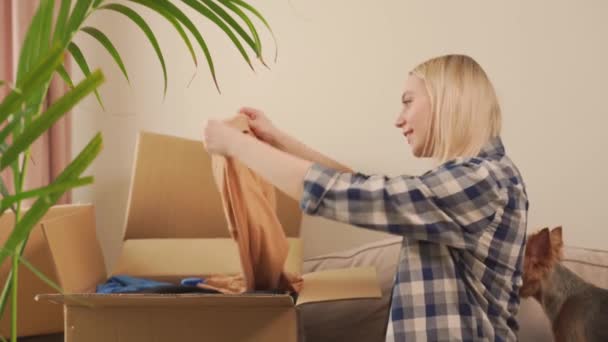 Kanepede oturan bir kadın taşındıktan sonra karton bir kutudan giysilerini çıkarıyor. — Stok video